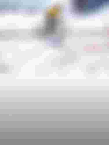 Cyclist in a yellow jacket and helmet rides a bike on a snowy road with mountains, village houses and trees in the background.