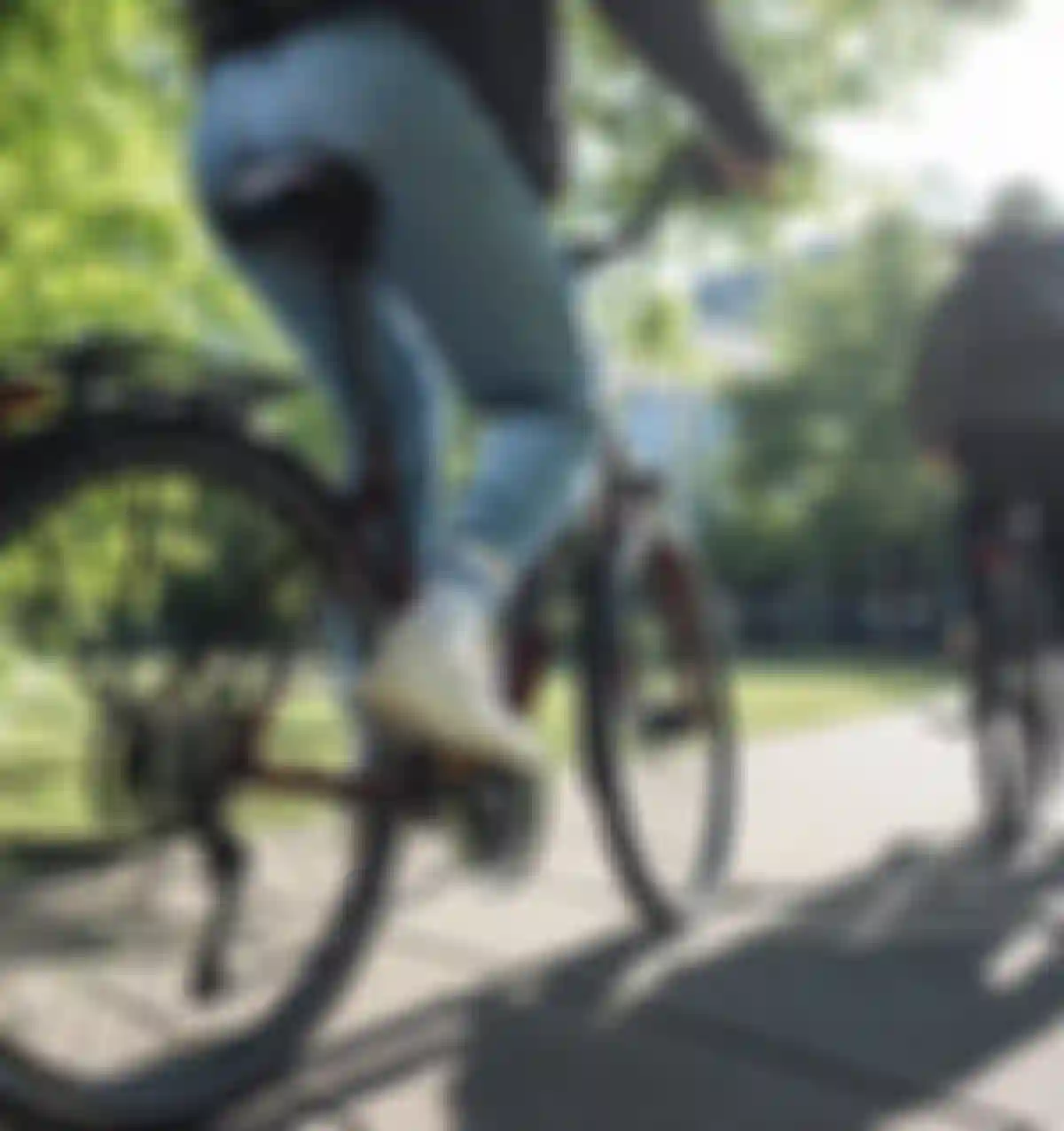 Two cyclists ride along a sunlit path in a park, surrounded by green trees and a modern building in the background.