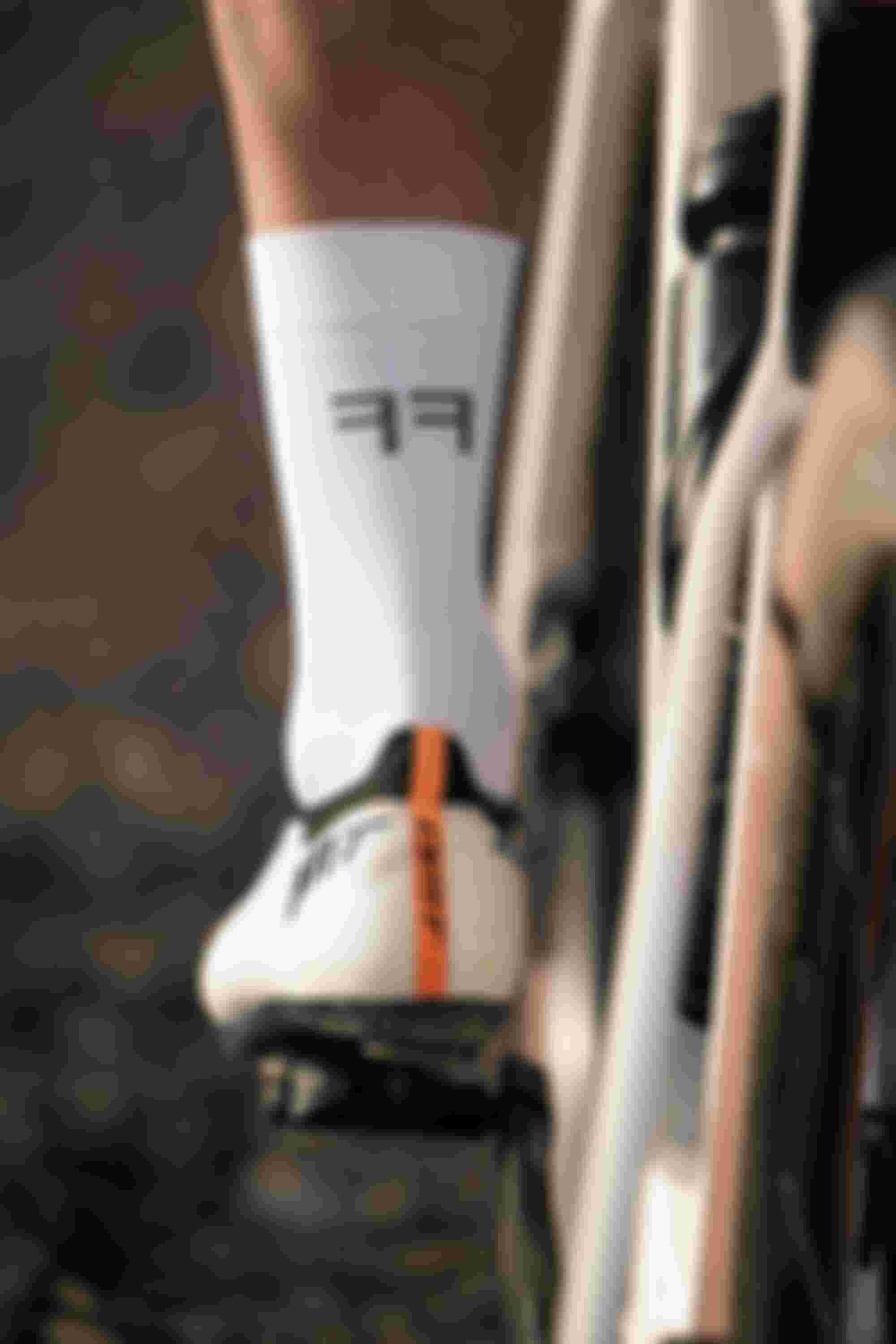 Close-up of a cyclist's foot on a pedal, wearing white socks with "FF" lettering and white cycling shoes with an orange stripe.