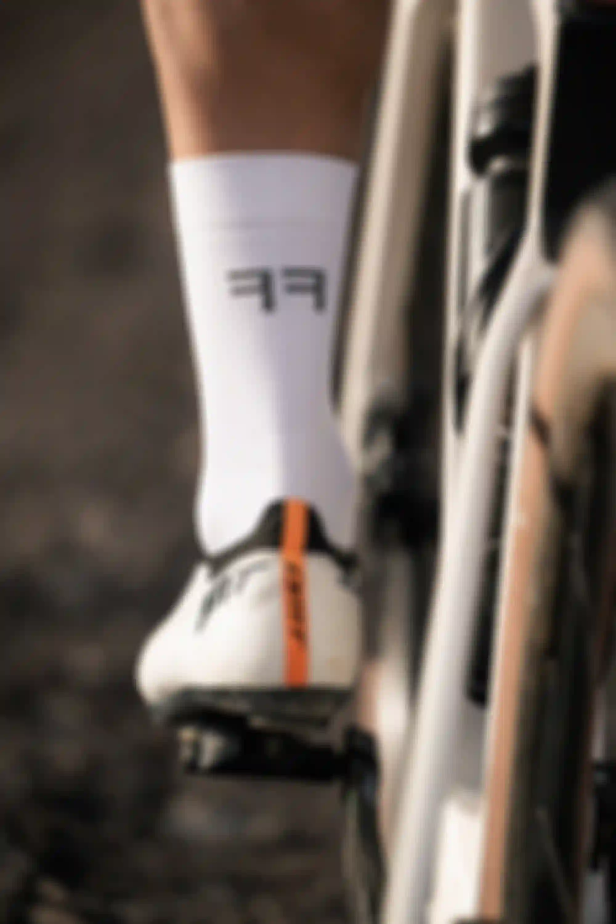 Close-up of a cyclist's foot on a pedal, wearing white socks with "FF" lettering and white cycling shoes with an orange stripe.