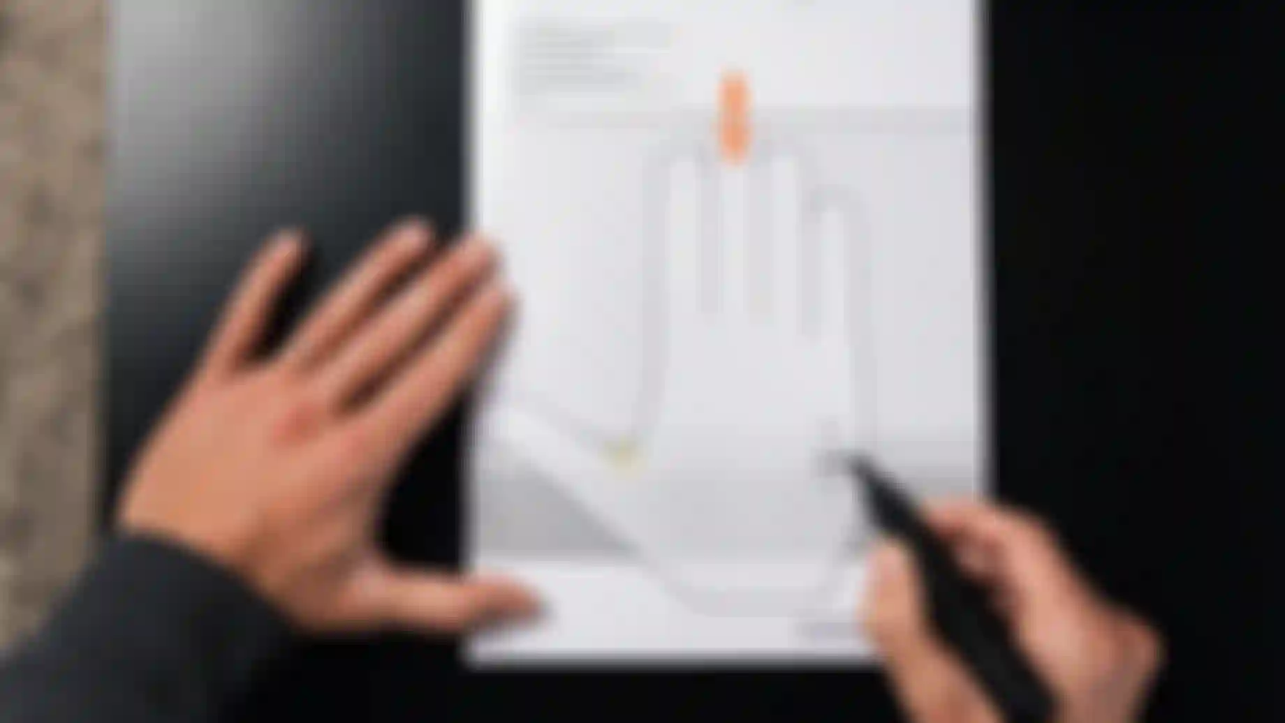 Person measuring hand size with a printed guide on a dark surface, using a marker to indicate dimensions on a diagram of a hand.