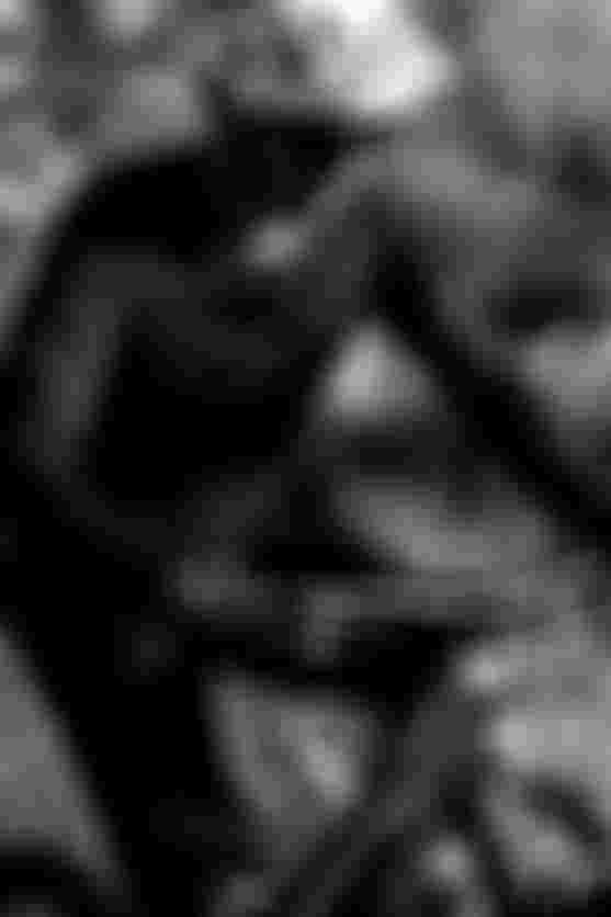 A cyclist wearing a helmet and sunglasses rides a mountain bike on a rugged trail, surrounded by blurred natural scenery. Black and white image.