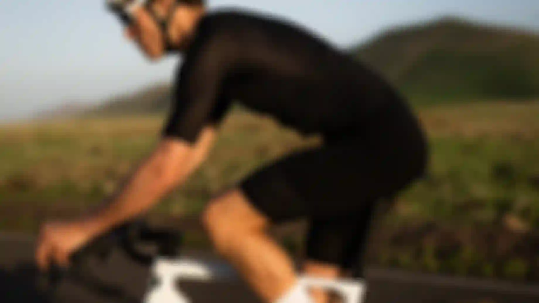 A cyclist wearing black gear and a white helmet rides a white bike on a road with grassy hills in the background.