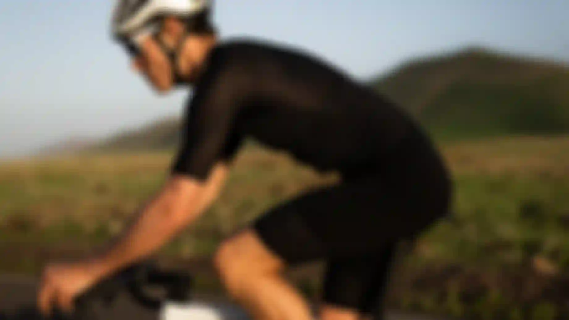 A cyclist wearing black gear and a white helmet rides a white bike on a road with grassy hills in the background.