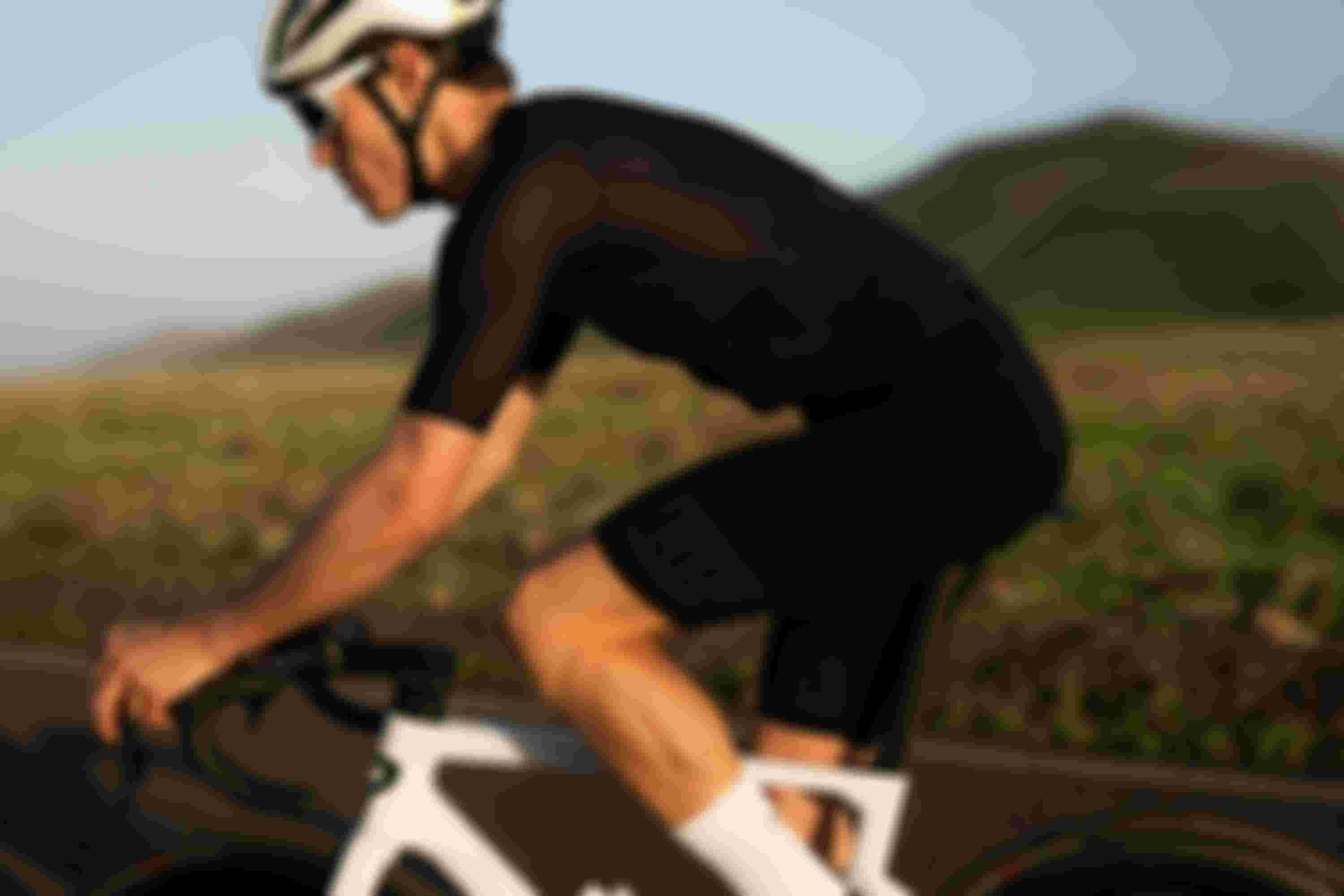 A cyclist wearing black gear and a white helmet rides a white bike on a road with grassy hills in the background.