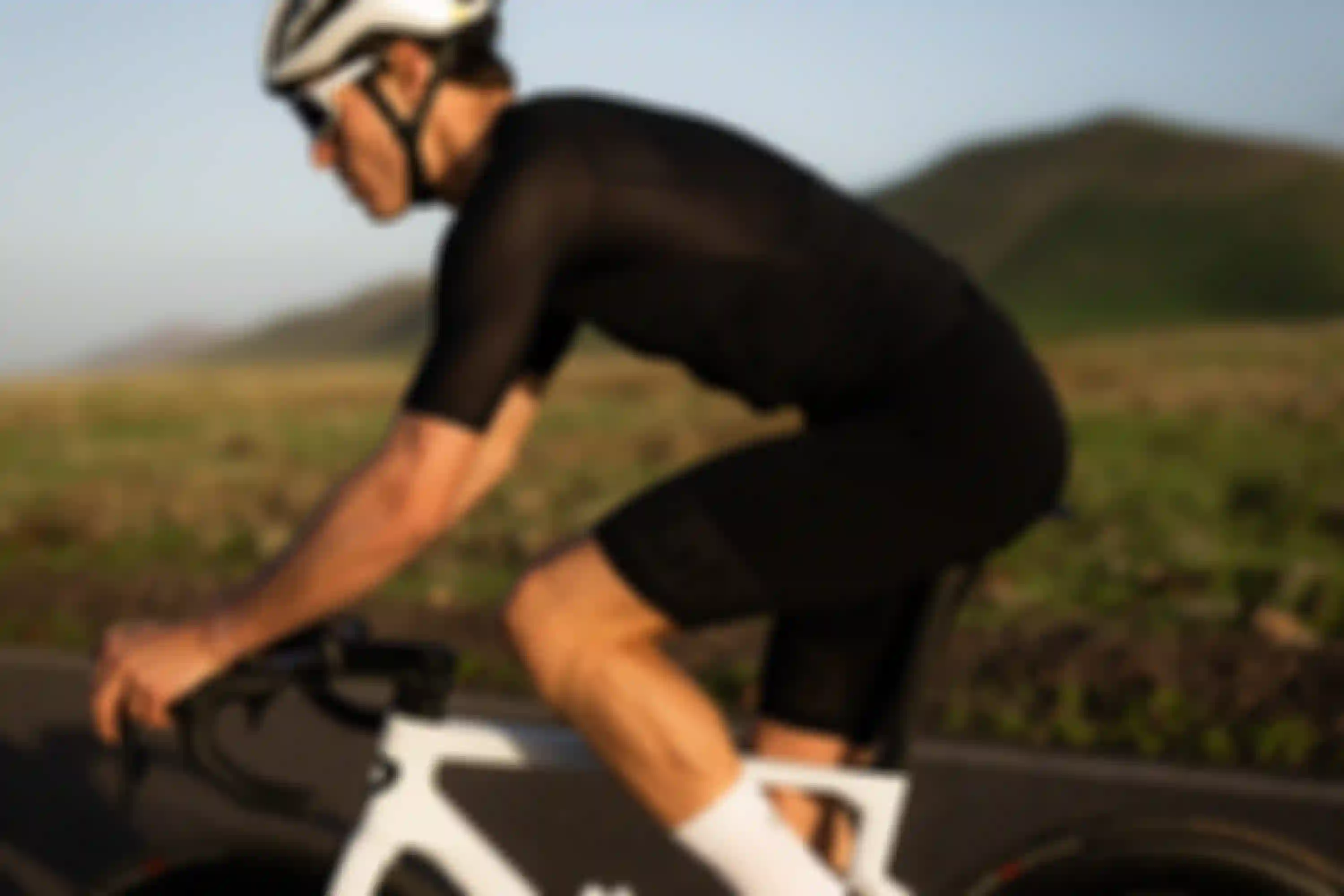 A cyclist wearing black gear and a white helmet rides a white bike on a road with grassy hills in the background.