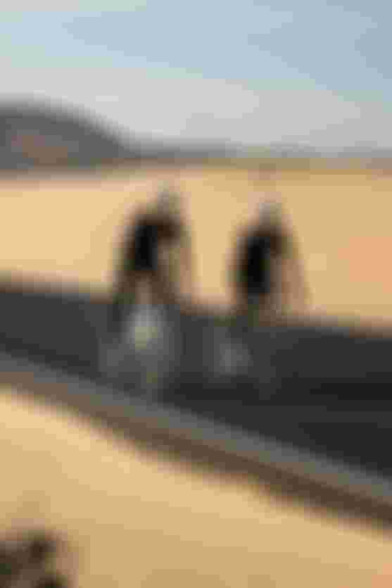 Two cyclists ride on a desert road, wearing matching black outfits and helmets, with a mountain in the background.