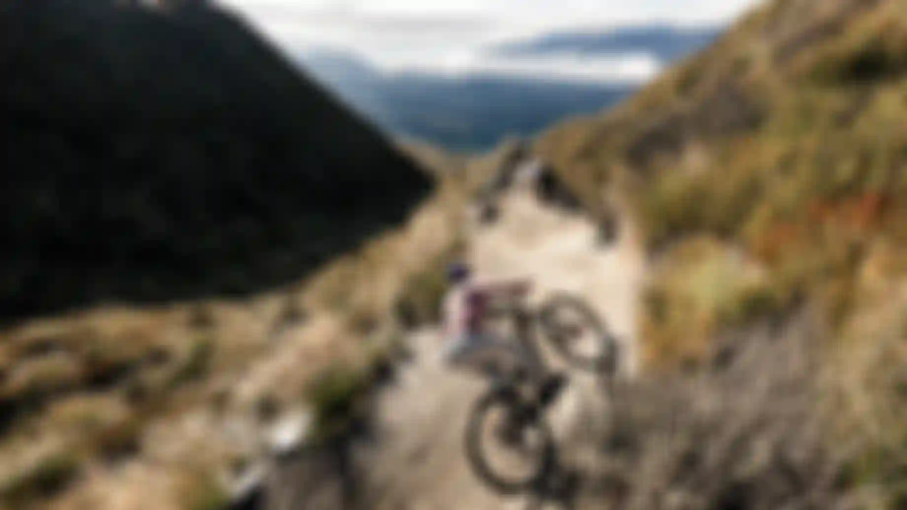 Two mountain bikers descending a flowing alpine singletrack trail, kicking up dust while cornering through a berm with wide mountain landscape in the background.