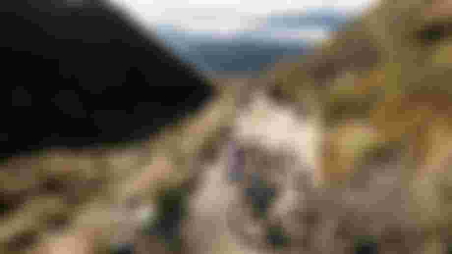 Two mountain bikers descending a flowing alpine singletrack trail, kicking up dust while cornering through a berm with wide mountain landscape in the background.