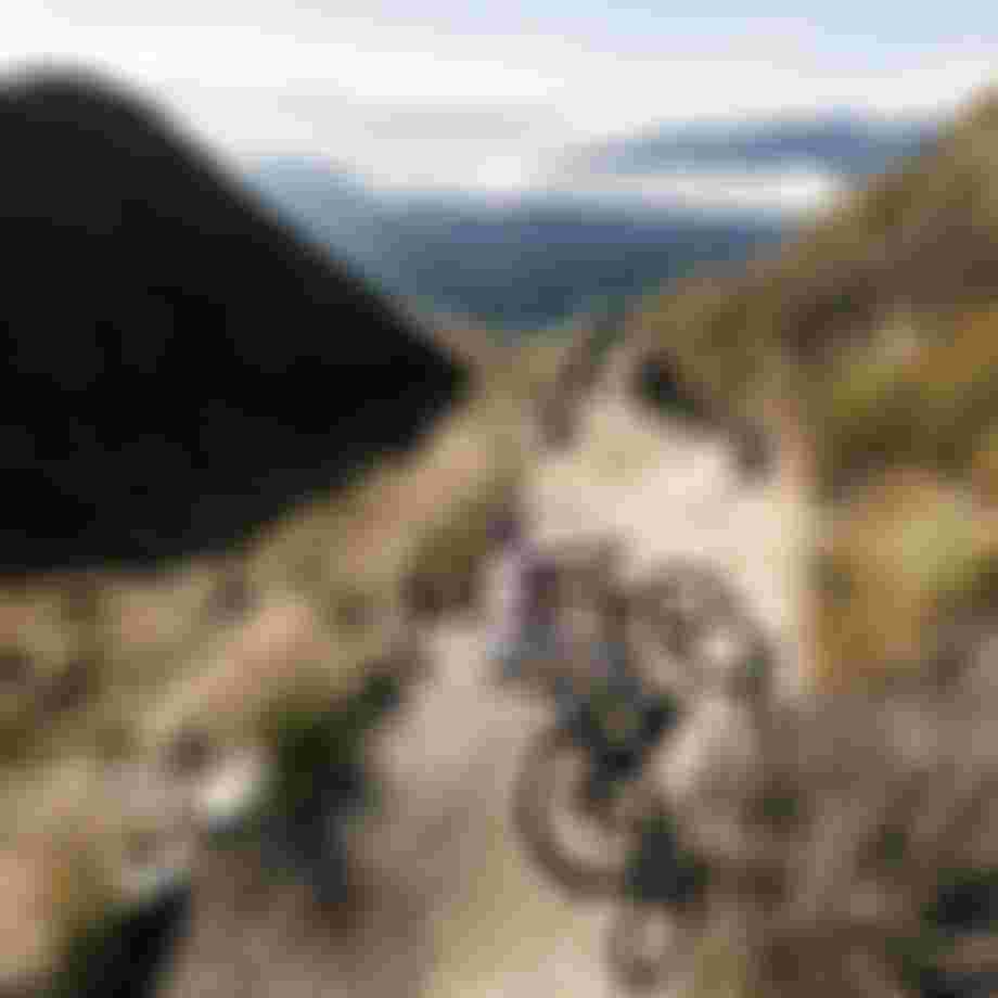 Two mountain bikers descending a flowing alpine singletrack trail, kicking up dust while cornering through a berm with wide mountain landscape in the background.