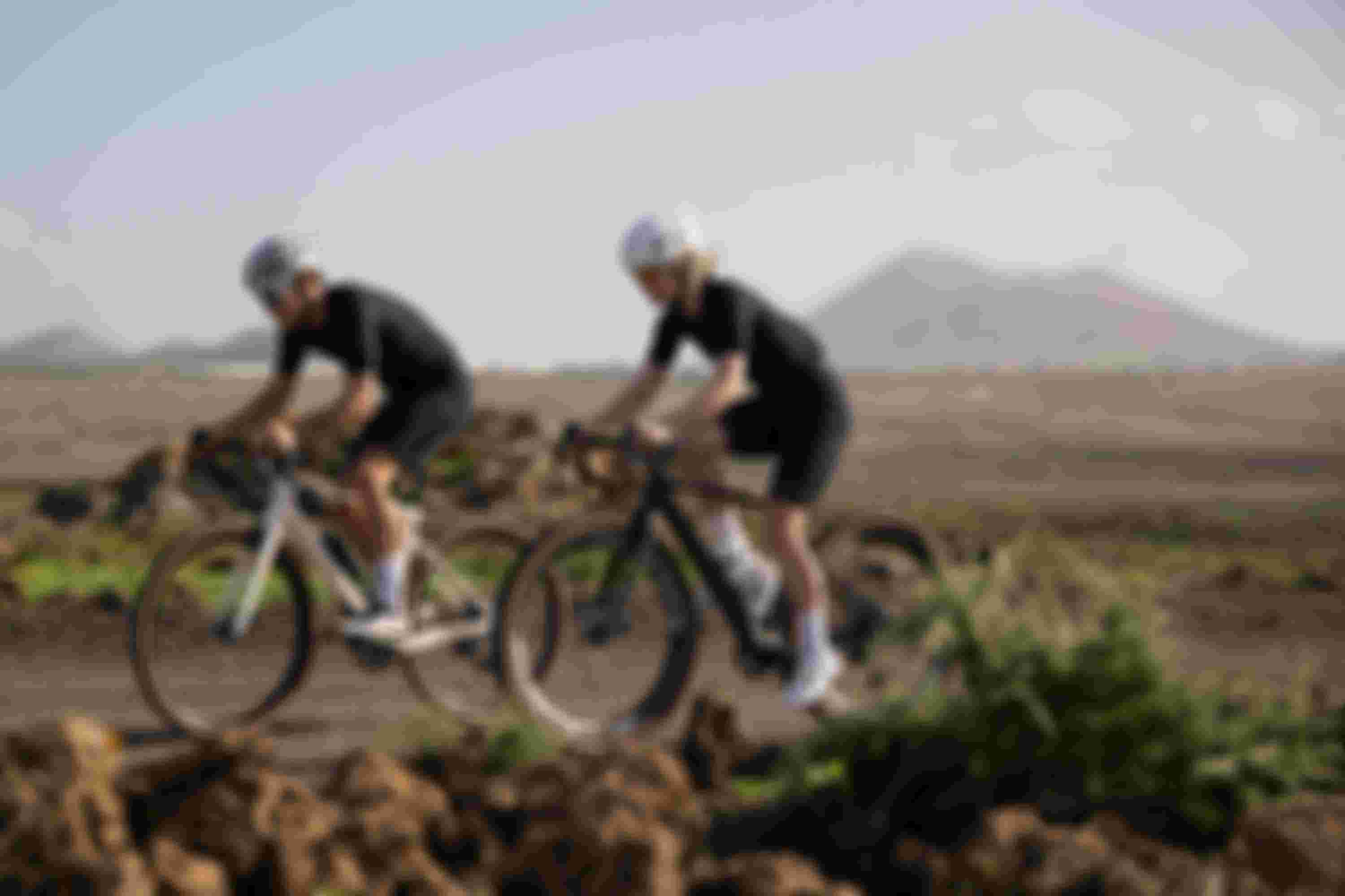 Two cyclists in black outfits and white helmets ride on a rocky path, with distant hills and a clear sky in the background.