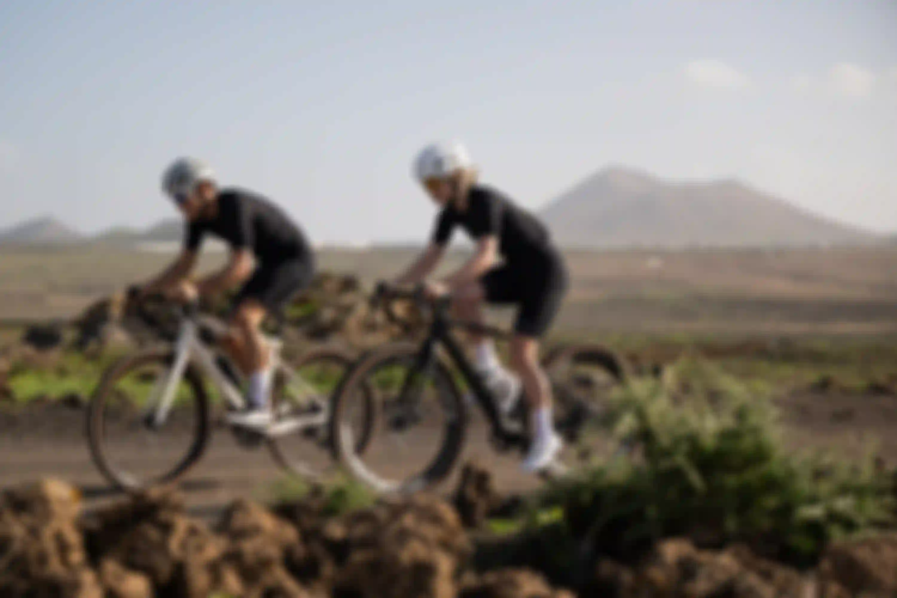 Two cyclists in black outfits and white helmets ride on a rocky path, with distant hills and a clear sky in the background.