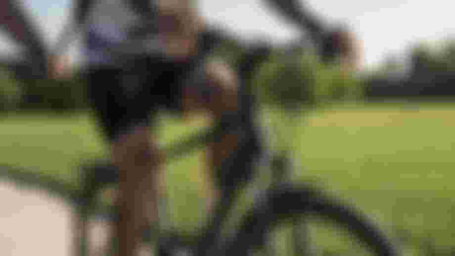 Close-up of a tattooed person riding a bicycle on a sunny day, with a lush green park in the background.