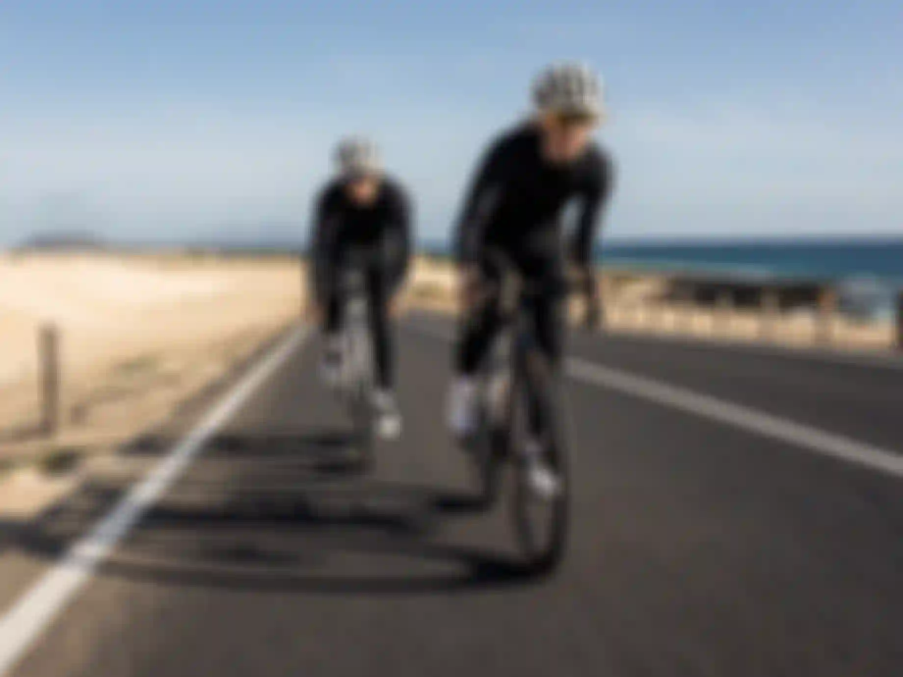 Two cyclists in black outfits ride on a coastal road, with sandy dunes on one side and the ocean on the other under a clear blue sky.
