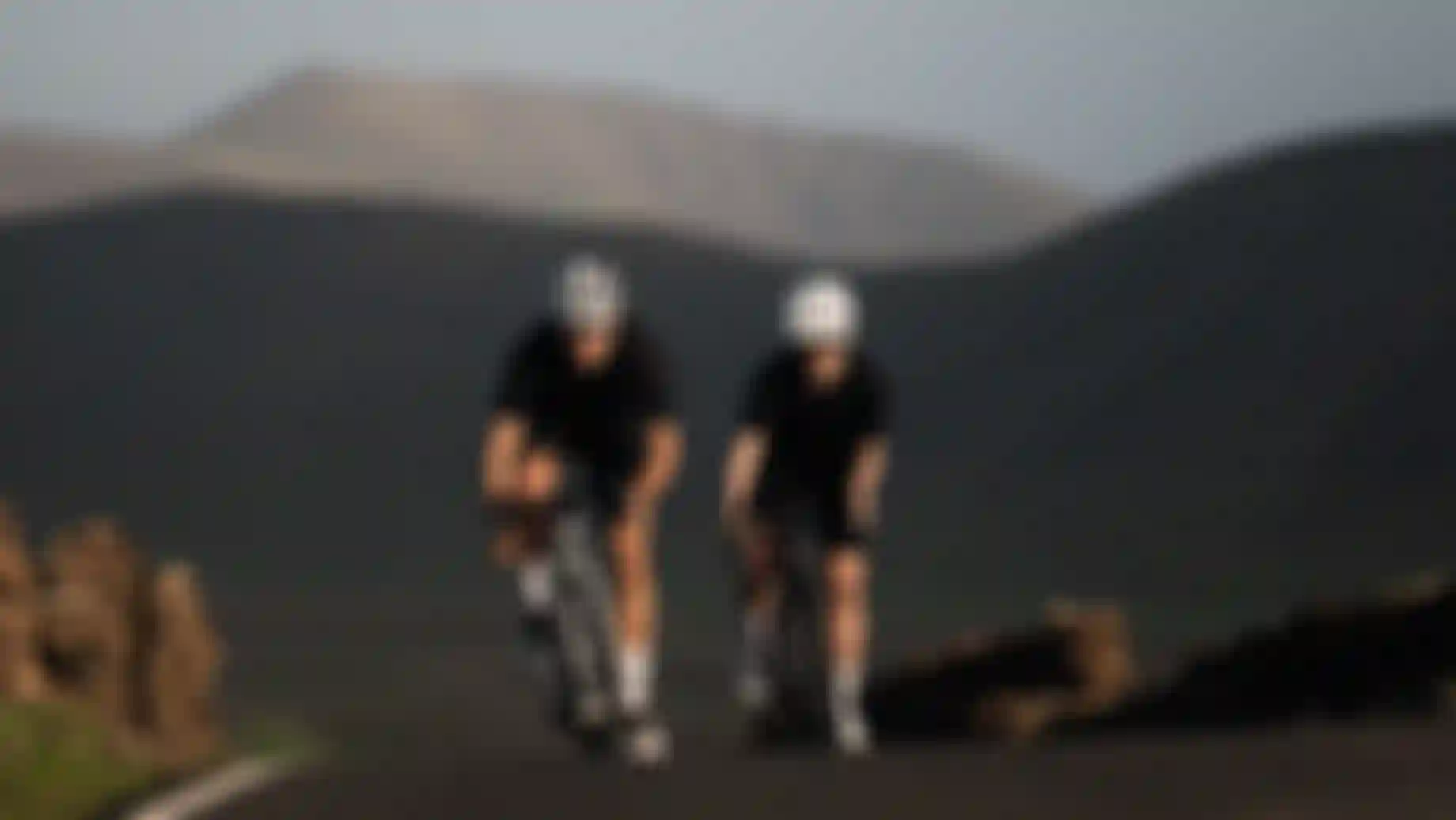 Two cyclists in black outfits ride uphill on a road with mountainous landscape in the background, under clear skies.