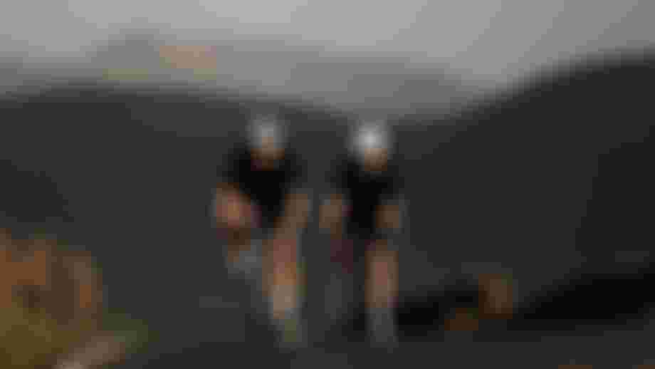 Two cyclists in black outfits ride uphill on a road with mountainous landscape in the background, under clear skies.