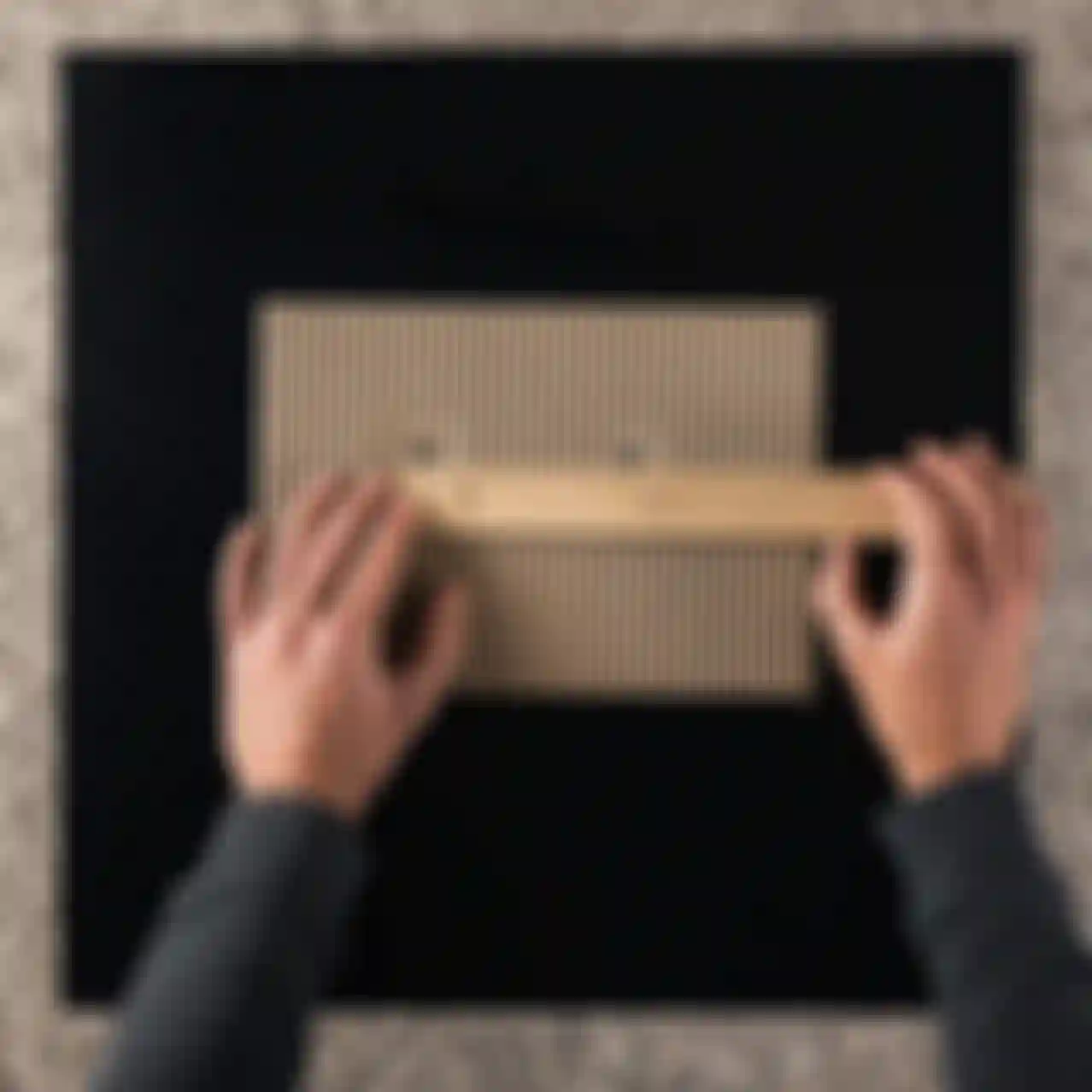 Hands measuring a corrugated cardboard sheet with a wooden ruler on a black tabletop. A black marker lies nearby.