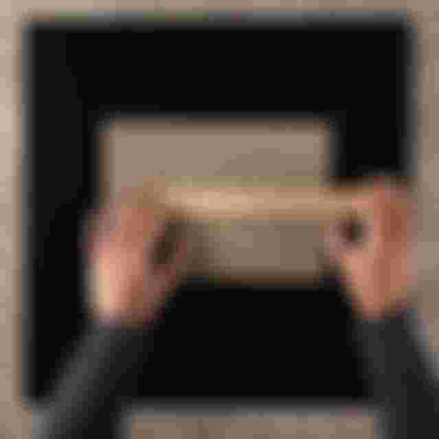 Hands measuring a corrugated cardboard sheet with a wooden ruler on a black tabletop. A black marker lies nearby.