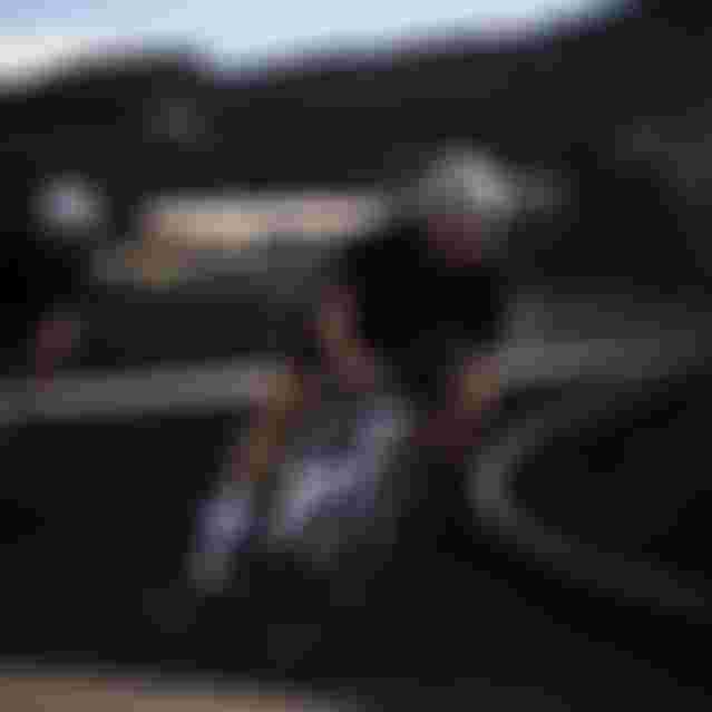 Two cyclists in black outfits and helmets speed down a winding road, leaning into a turn, under a clear blue sky.
