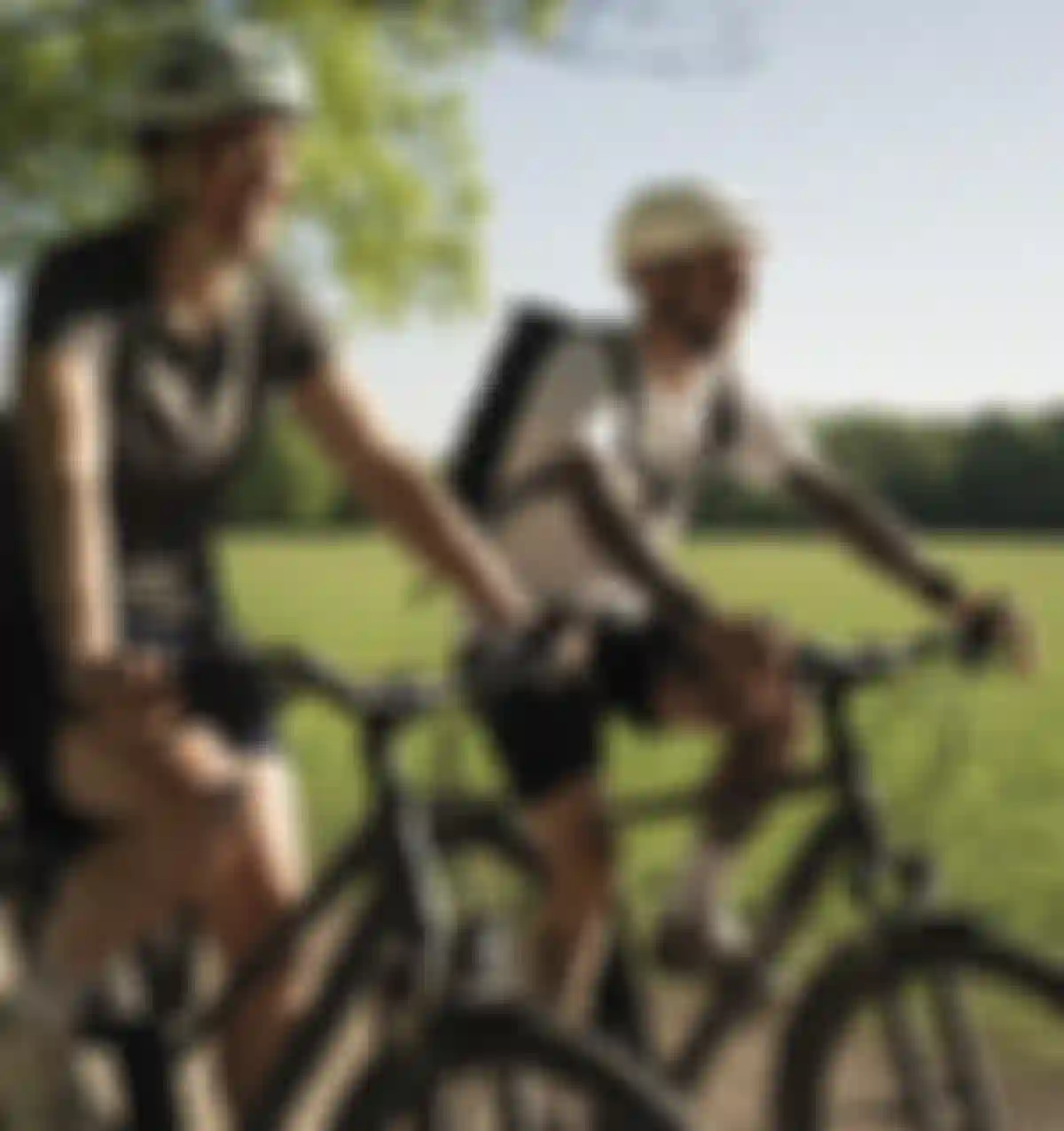 Two people biking on a sunny path in a park, wearing helmets and casual clothes, with green grass and trees in the background.