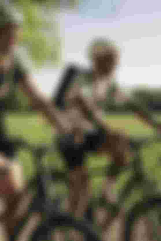 Two people biking on a sunny path in a park, wearing helmets and casual clothes, with green grass and trees in the background.