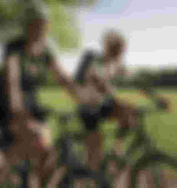 Two people biking on a sunny path in a park, wearing helmets and casual clothes, with green grass and trees in the background.