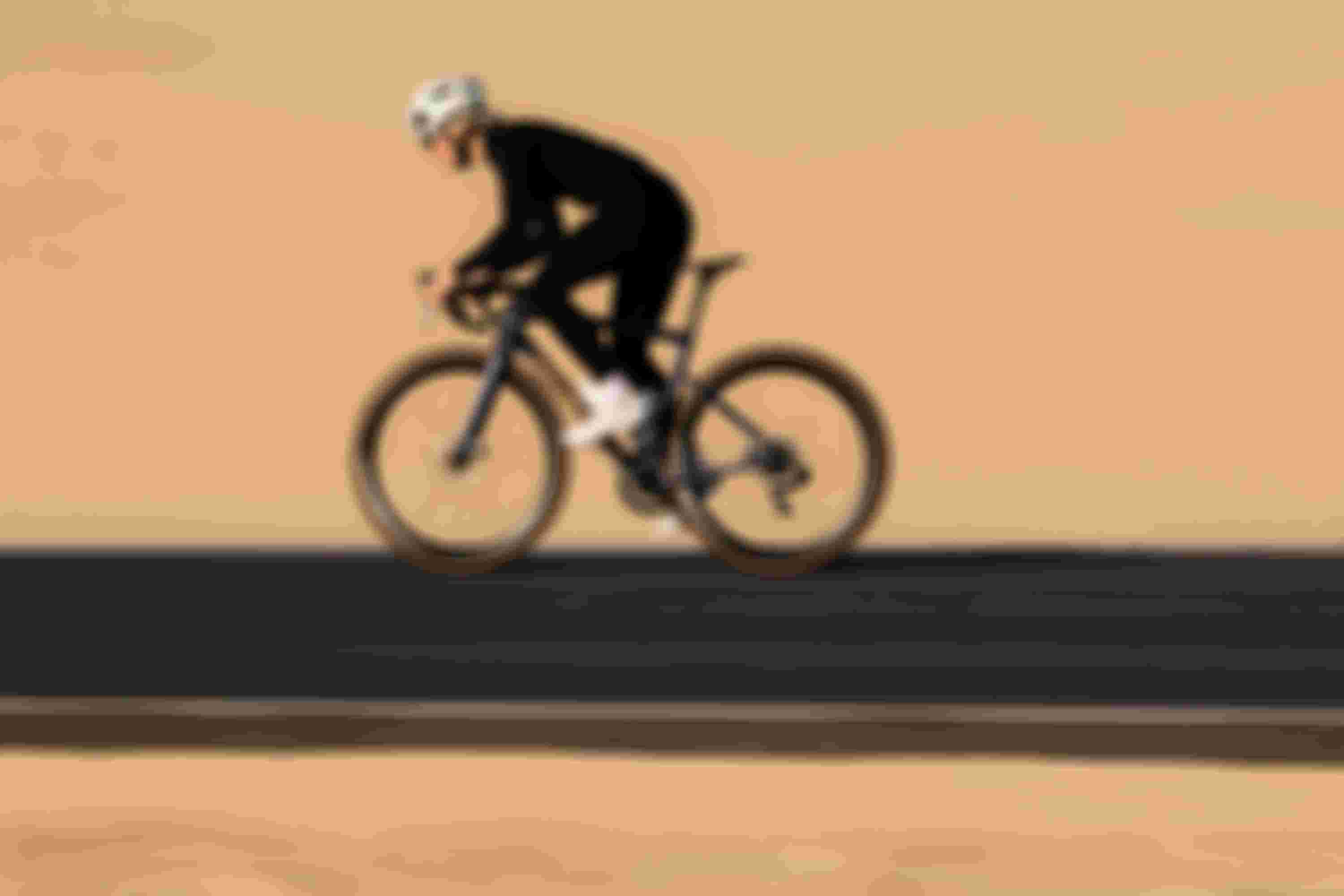 A cyclist wearing a helmet and black attire rides a sleek bicycle on a smooth road through a desert landscape.
