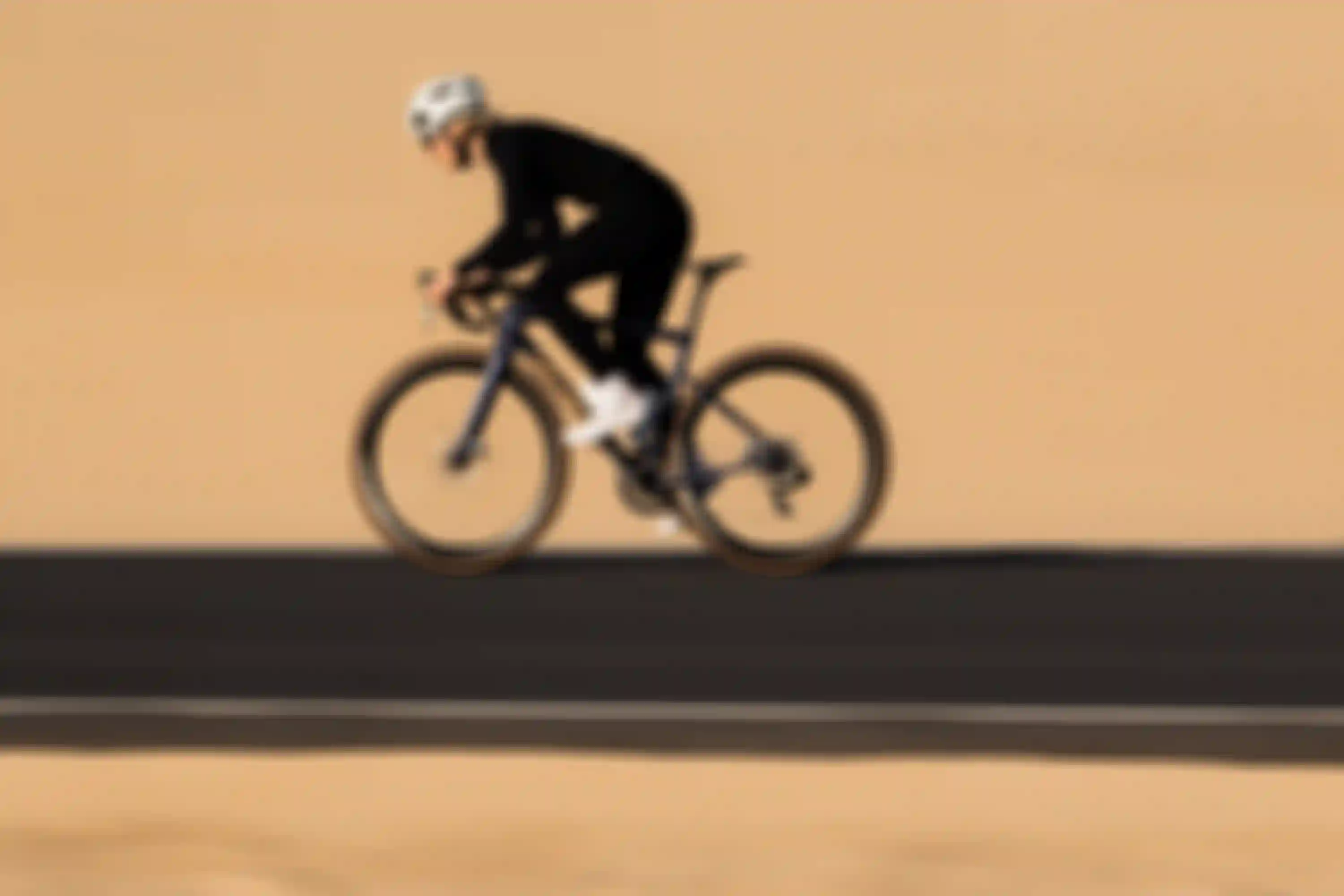 A cyclist wearing a helmet and black attire rides a sleek bicycle on a smooth road through a desert landscape.