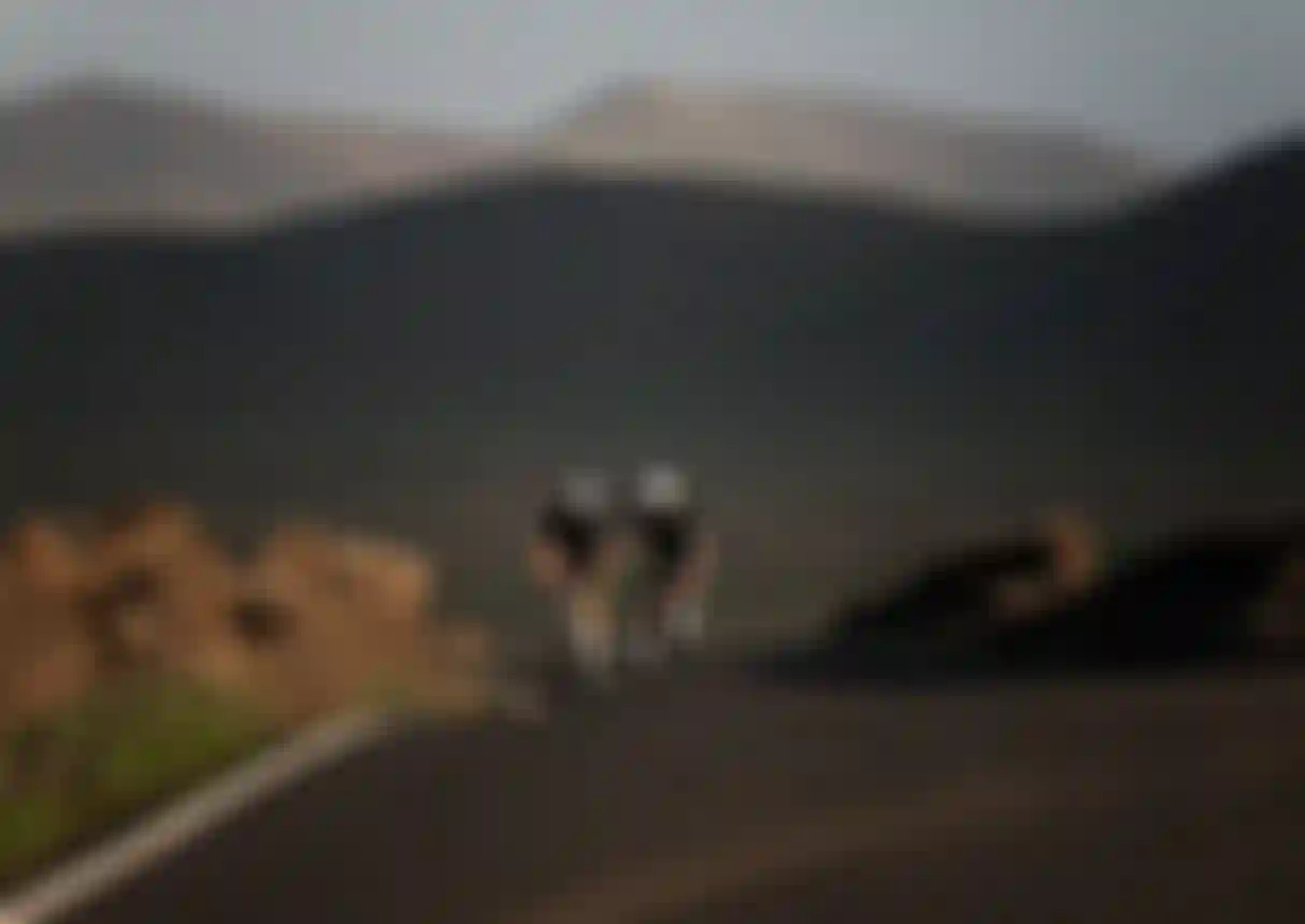Two cyclists wearing helmets ride up a winding road through a rocky, mountainous landscape under a clear sky.