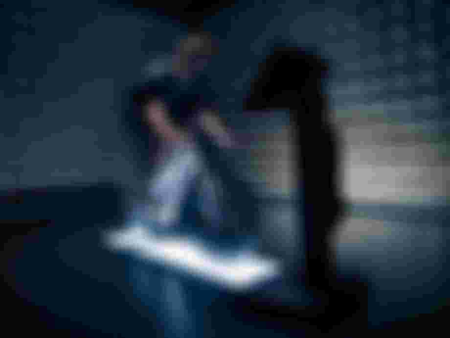 Cyclist wearing a mask rides a stationary bike in a dimly lit room with soundproof walls and a monitor in front.