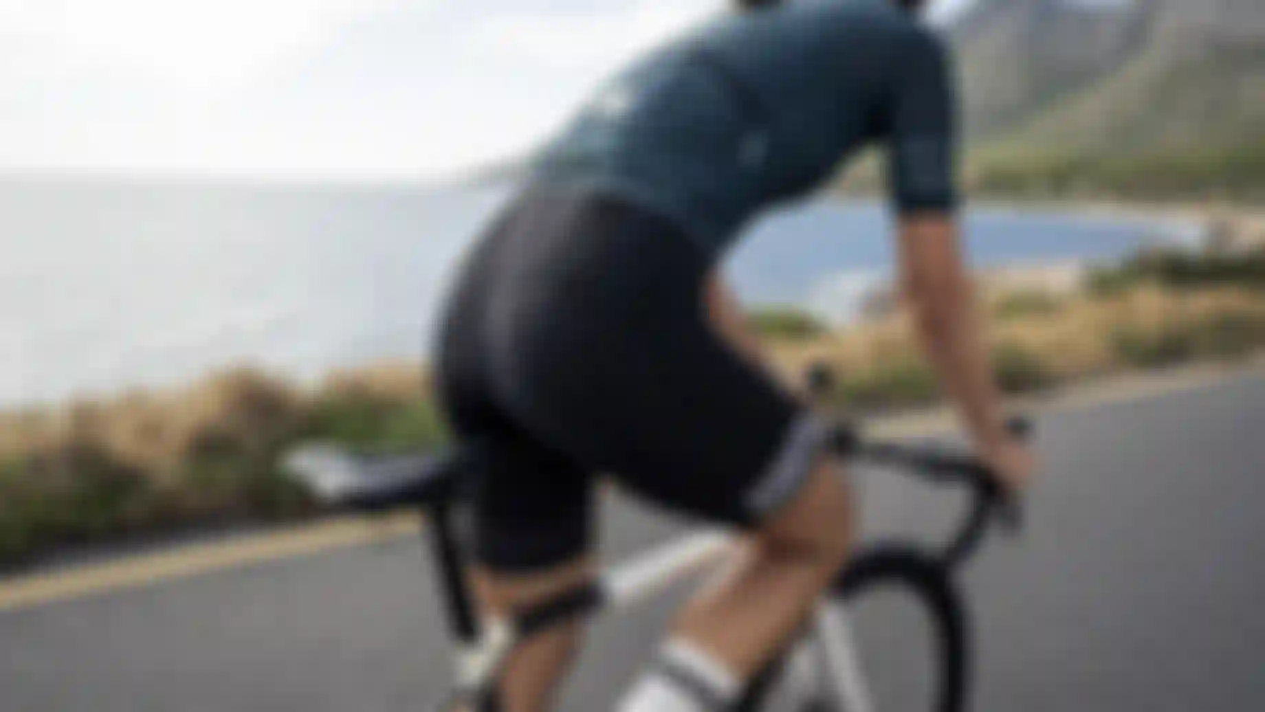 Person fährt auf einem Rennrad entlang einer Küstenstraße mit Blick auf das Meer und Berge im Hintergrund. Die Person trägt ein enges Radtrikot und eine gepolsterte Radhose.