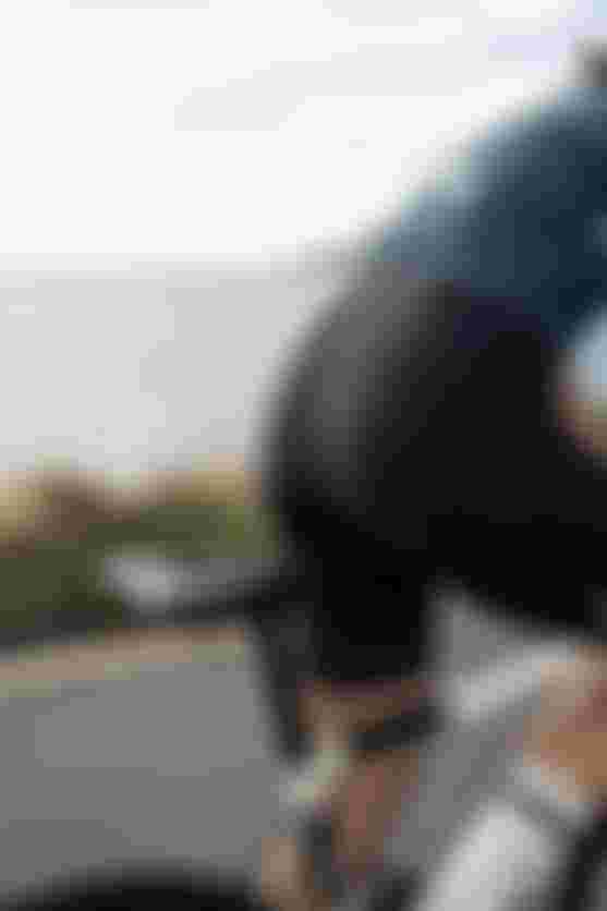 Person fährt auf einem Rennrad entlang einer Küstenstraße mit Blick auf das Meer und Berge im Hintergrund. Die Person trägt ein enges Radtrikot und eine gepolsterte Radhose.