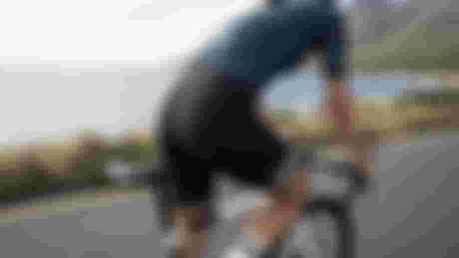 Person fährt auf einem Rennrad entlang einer Küstenstraße mit Blick auf das Meer und Berge im Hintergrund. Die Person trägt ein enges Radtrikot und eine gepolsterte Radhose.