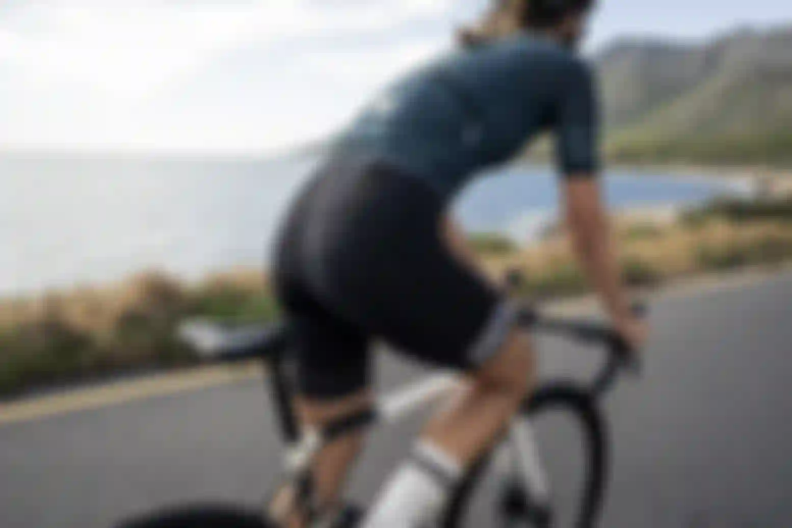 Person fährt auf einem Rennrad entlang einer Küstenstraße mit Blick auf das Meer und Berge im Hintergrund. Die Person trägt ein enges Radtrikot und eine gepolsterte Radhose.