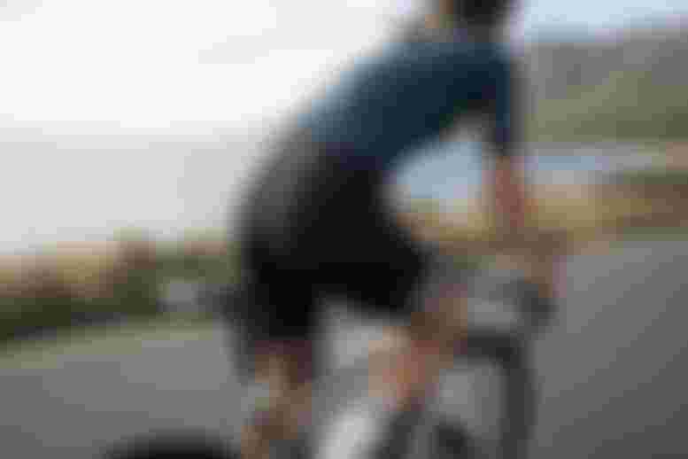 Person fährt auf einem Rennrad entlang einer Küstenstraße mit Blick auf das Meer und Berge im Hintergrund. Die Person trägt ein enges Radtrikot und eine gepolsterte Radhose.