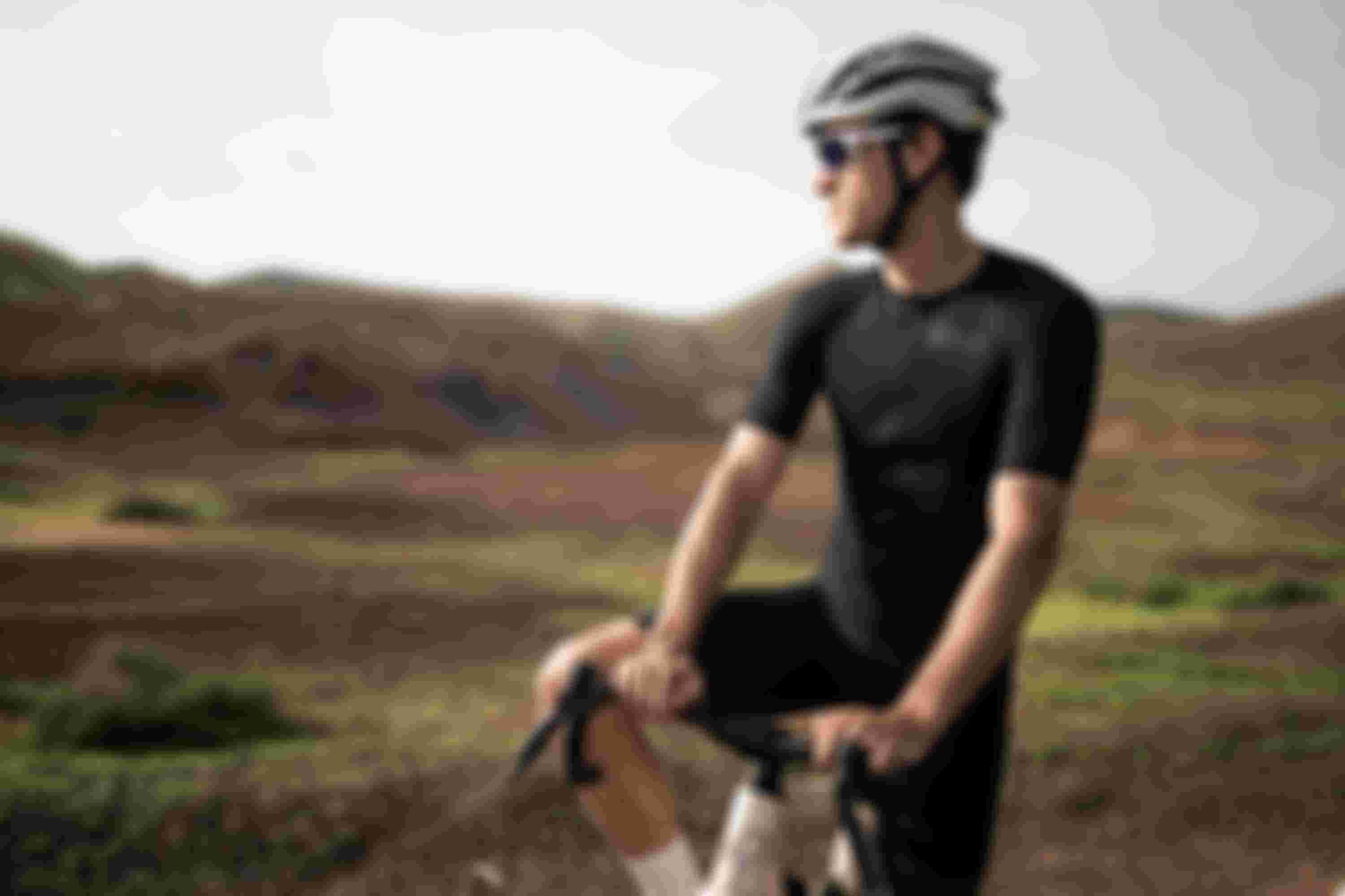 A cyclist wearing a black outfit and helmet, resting on a bike in a scenic, arid landscape with hills in the background.