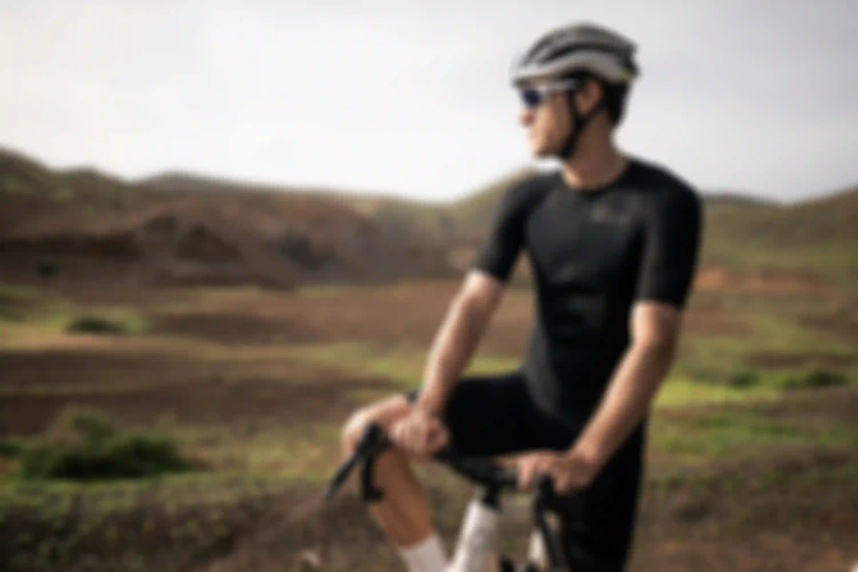 A cyclist wearing a black outfit and helmet, resting on a bike in a scenic, arid landscape with hills in the background.