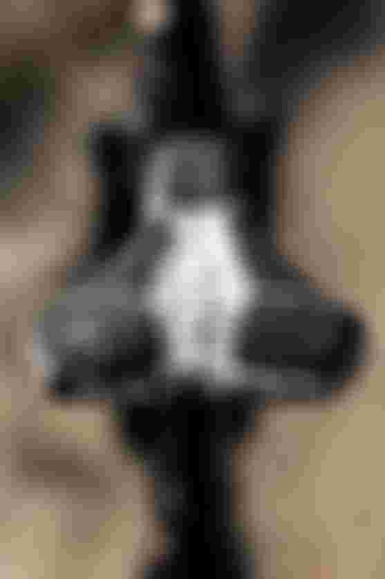 Close-up of a bicycle seat with a black and white speckled pattern, featuring a small logo on the front, set against a blurred outdoor background.