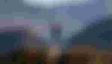 A mountain biker rides down a trail, arms outstretched, against a backdrop of rugged mountains and a distant lake under a clear sky.