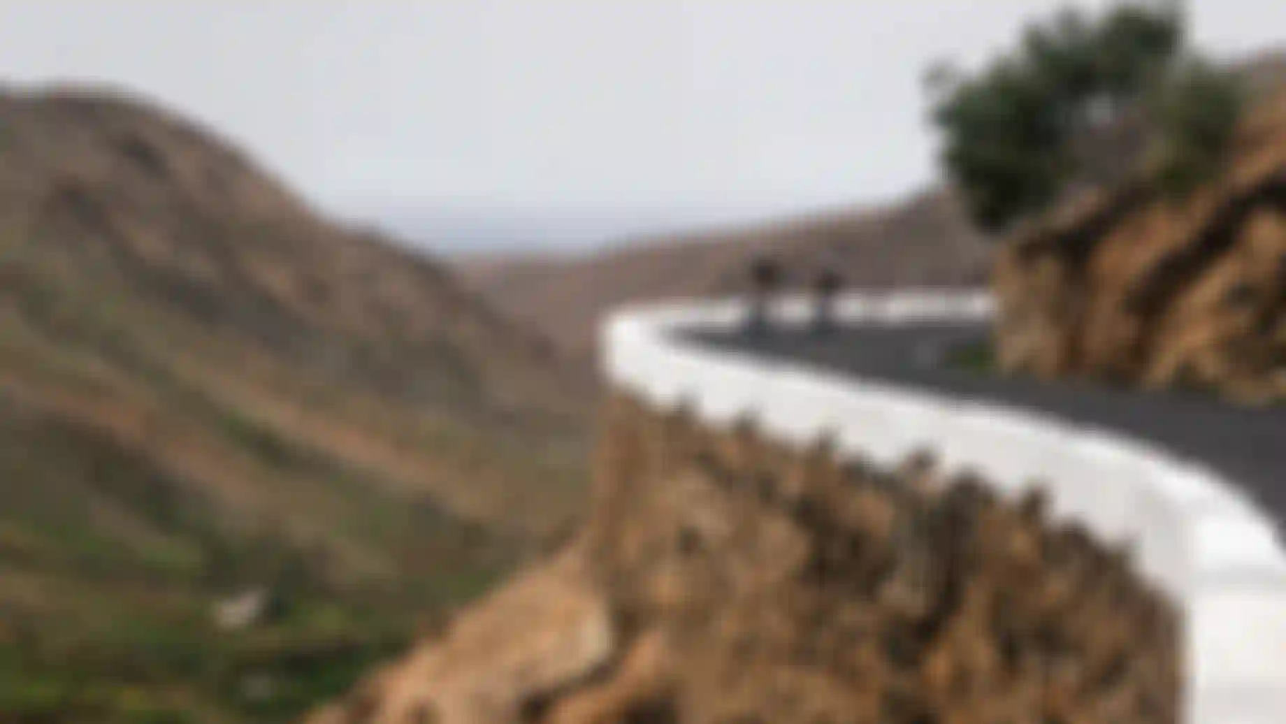 Two cyclists ride on a mountain road curve with a stone wall, surrounded by rugged hills and a distant view of green fields.