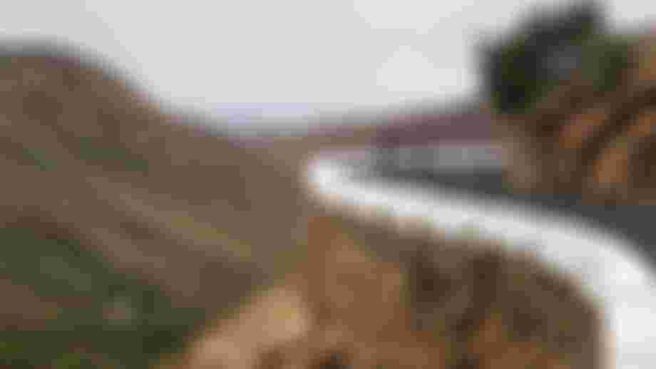 Two cyclists ride on a mountain road curve with a stone wall, surrounded by rugged hills and a distant view of green fields.