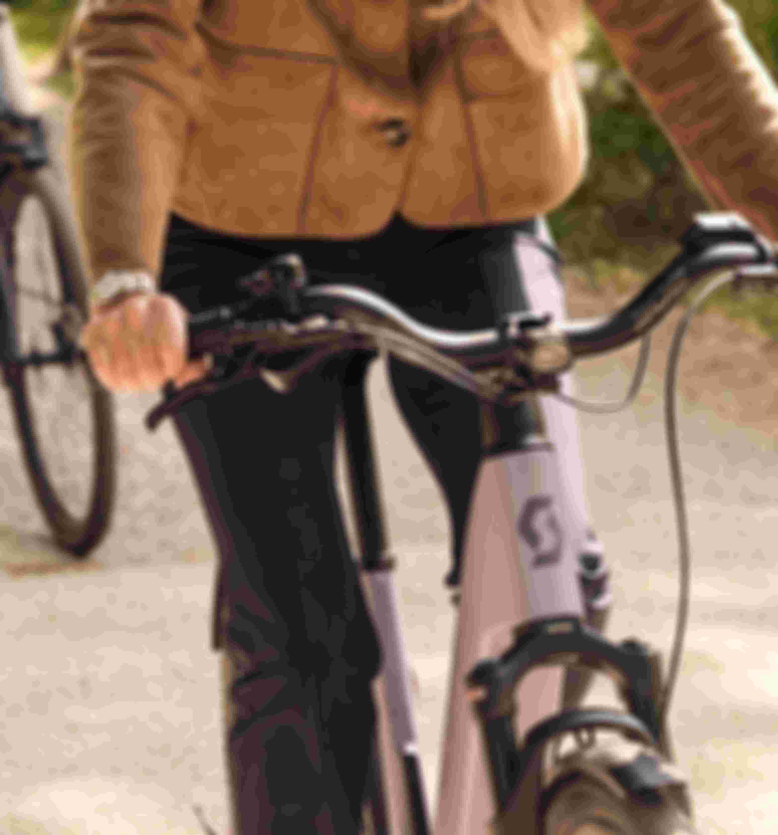 A person in a tan jacket and dark pants rides a purple Scott bike on a sunny path, holding the handlebars firmly.