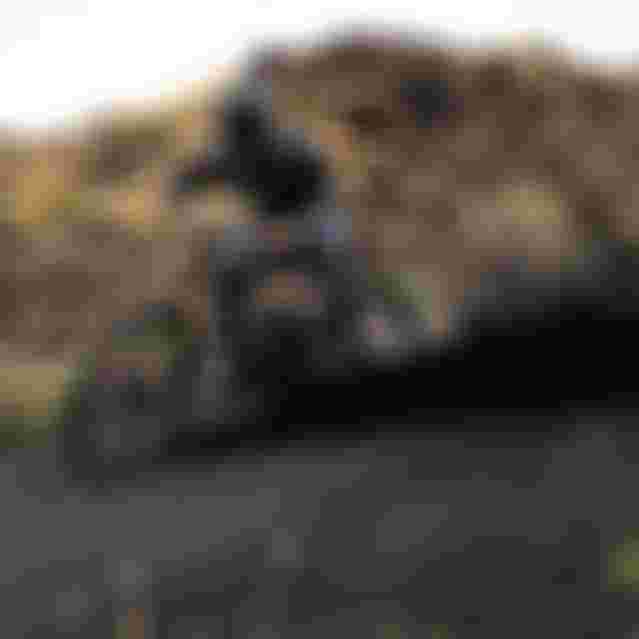 Mountain biker in black gear skids dramatically on a dirt trail, creating a plume of dust against a rugged, rocky landscape.