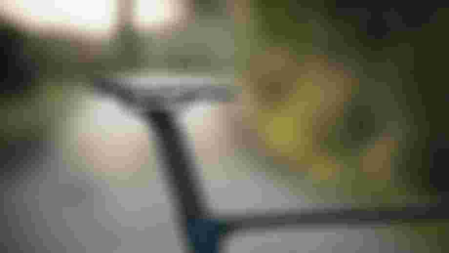 Close-up photo of a road bike saddle and seatpost with water droplets, set against a blurred outdoor road and green background in soft evening light.