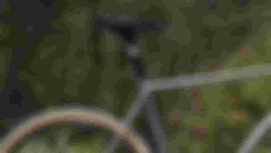 Side-view product shot of a gravel bike with a grey frame, drop handlebars, disc brakes, and tan-wall tires, standing on a gravel road with blurred green foliage in the background.
