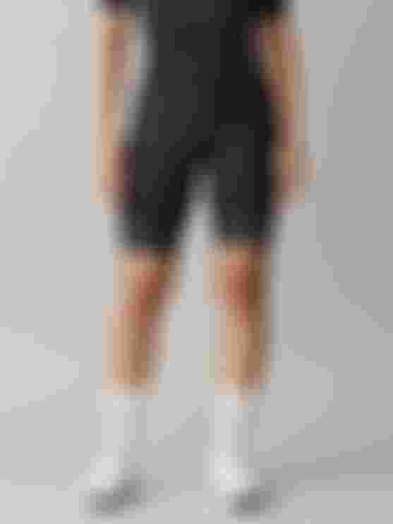 Female cyclist wearing black SQlab ONE12 shorts, standing front-facing, with a layered 3D cutaway of the pad construction shown in front of her.
