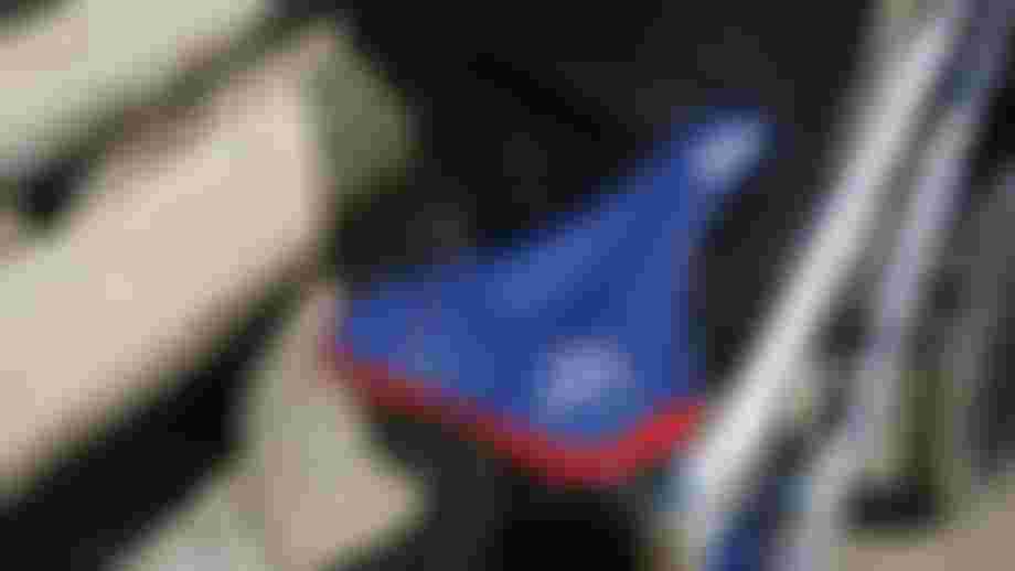 Blue SQlab bicycle saddle with red trim and geometric graphics, mounted on a road bike. The saddle is photographed from a slight overhead angle, with parts of the bike frame and a stone surface visible in the background.