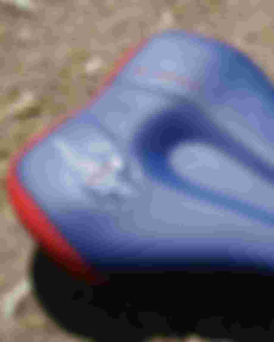 Blue SQlab bicycle saddle with red trim and geometric graphics, mounted on a road bike. The saddle is photographed from a slight overhead angle, with parts of the bike frame and a stone surface visible in the background.