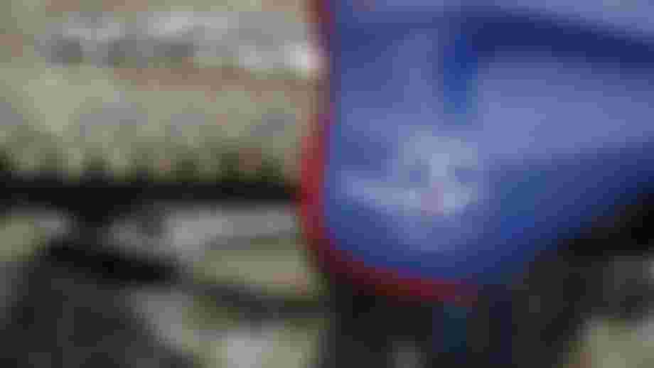 Blue SQlab bicycle saddle with red trim and geometric graphics, mounted on a road bike. The saddle is photographed from a slight overhead angle, with parts of the bike frame and a stone surface visible in the background.