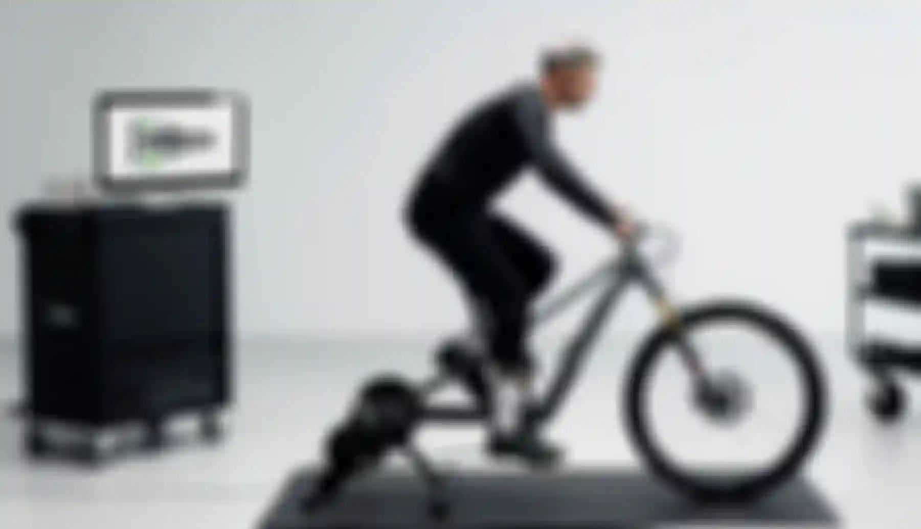 Male cyclist riding a mountain bike on an indoor trainer in a lab setting, with an SQlab saddle displayed on a monitor in the background.